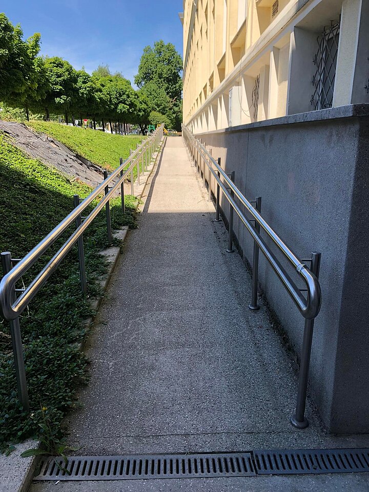 Pochylnia z poręczami prowadząca wzdłuż budynku, umożliwiająca dostęp osobom poruszającym się na wózkach