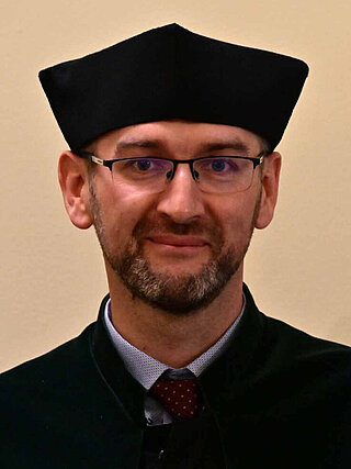 Oficjalna fotografia portretowa dr. inż. Tomasza Włodka, Prodziekana ds. Kształcenia Wydziału Wiertnictwa, Nafty i Gazu AGH. Sylwetka ujęta od pasa w górę, w czarnej todze akademickiej z biretem, na jednolitym jasnym tle studyjnym.
