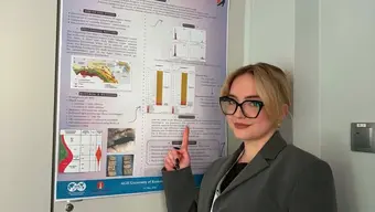 Zdjęcie przedstawia Studentkę WWNiG przy posterze z własnym artykułem na sesji naukowej. Studentka ubrana w szarą marynarkę, widok lewego profilu do pasa