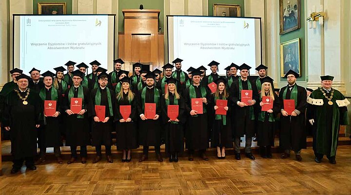 Społeczność studencka WNiG AGH Studentki i studenci WNiG AGH w trakcie rozdania dyplomów w Auli Głównej AGH. ustawieni w 3 szeregach jeden za drugim, wszyscy ubrani w odświętne togi, większość trzyma czerwone oprawki dyplomów ukończenia WWNiG